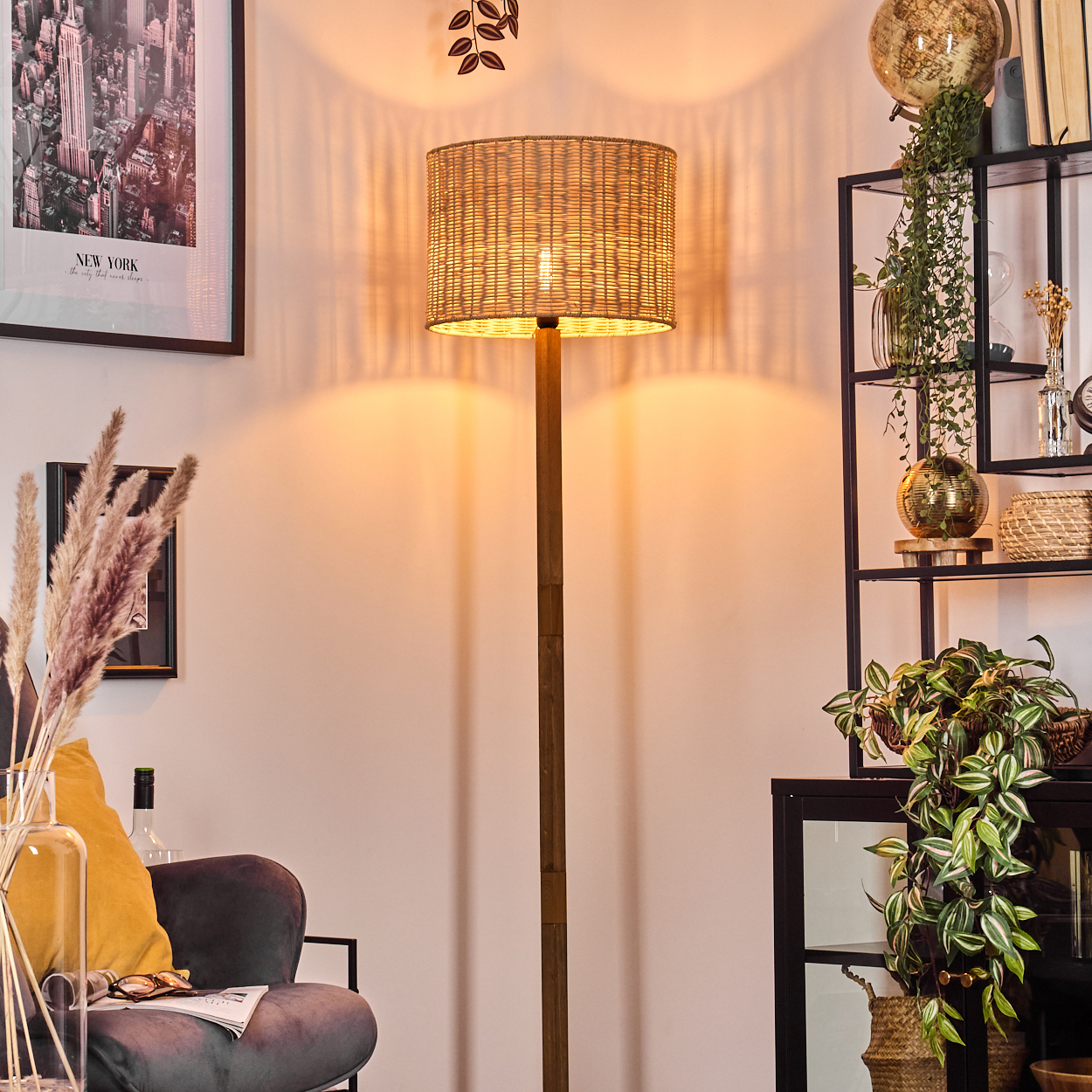 Eine beleuchtete Stehlampe mit gewebtem Schirm steht in einem gemütlichen Raum. Rechts ein Regal mit Deko und Pflanzen, an der Wand ein New York-Druck.