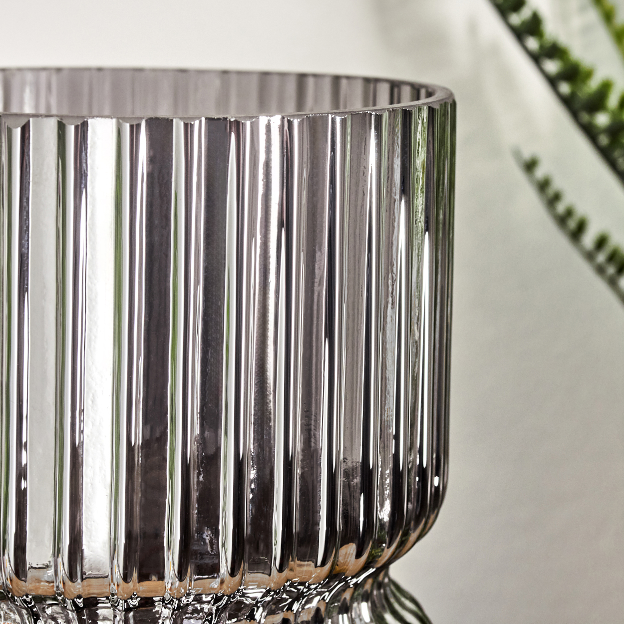 Nahaufnahme einer glänzenden, gerippten Glasvase. Die Vase reflektiert Licht. Grüne Pflanze im Hintergrund.
