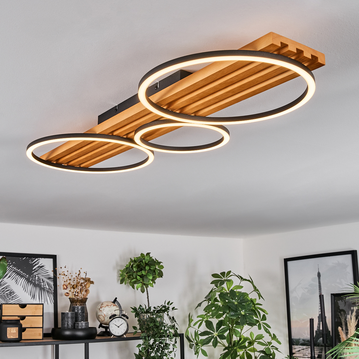 Moderne Deckenlampe mit drei kreisförmigen Lichtern. Der Hintergrund zeigt ein Wohnzimmer mit Pflanzen, gerahmten Bildern und einem Regal.