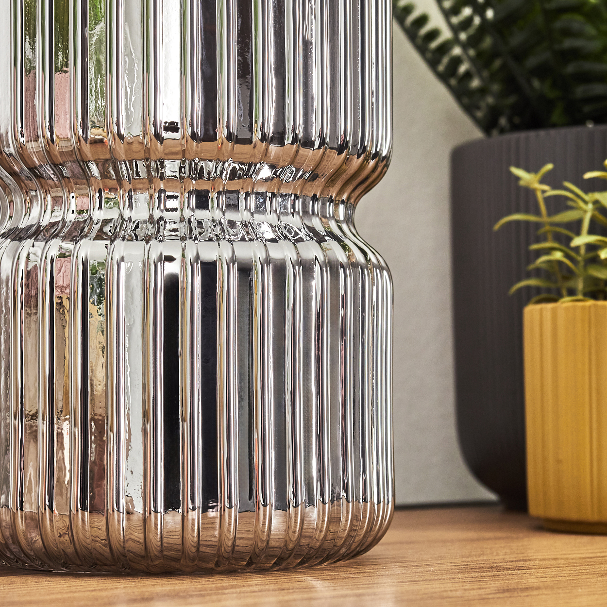 Eine glänzende, silberne Vase mit vertikalen Rippen. Im Hintergrund ein dunkelgrauer Topf und ein kleiner gelber Topf mit Pflanzen.