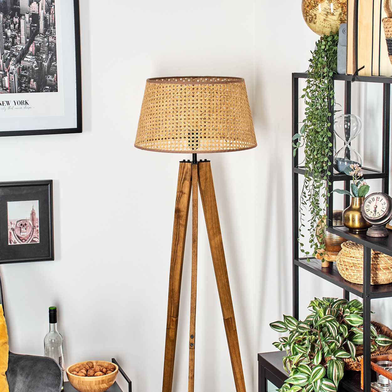 Eine Stehlampe mit Holzstativ und gewebtem Schirm steht in einem Raum mit Regal und Dekoration, darunter Pflanzen und ein Globus.