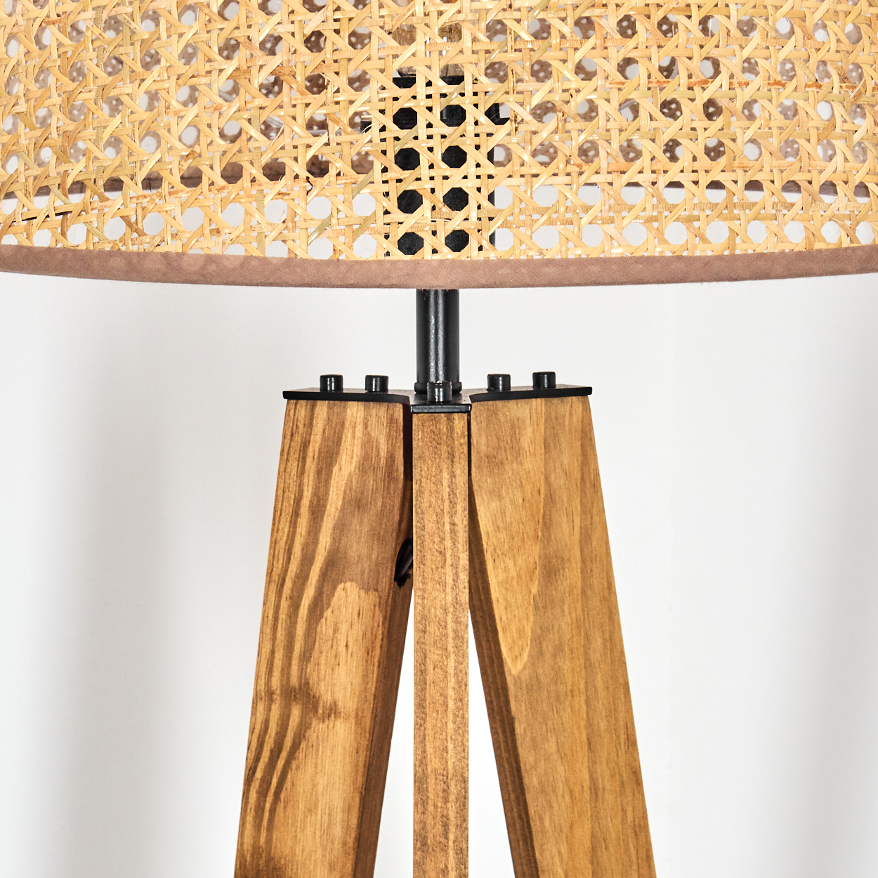 Nahaufnahme einer Lampe. Sie hat einen gewebten Lampenschirm, Holzbeine und einen schwarzen Metallrahmen. Der Hintergrund ist weiß.