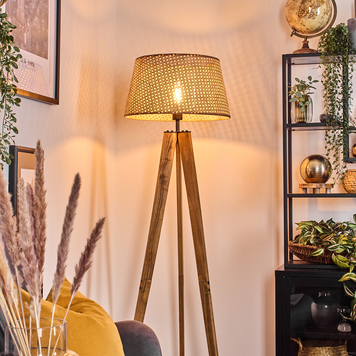 Eine hölzerne Stehlampe mit gewebtem Schirm beleuchtet einen gemütlichen Raum. Pflanzen, ein Globus und ein Regal befinden sich im Hintergrund.