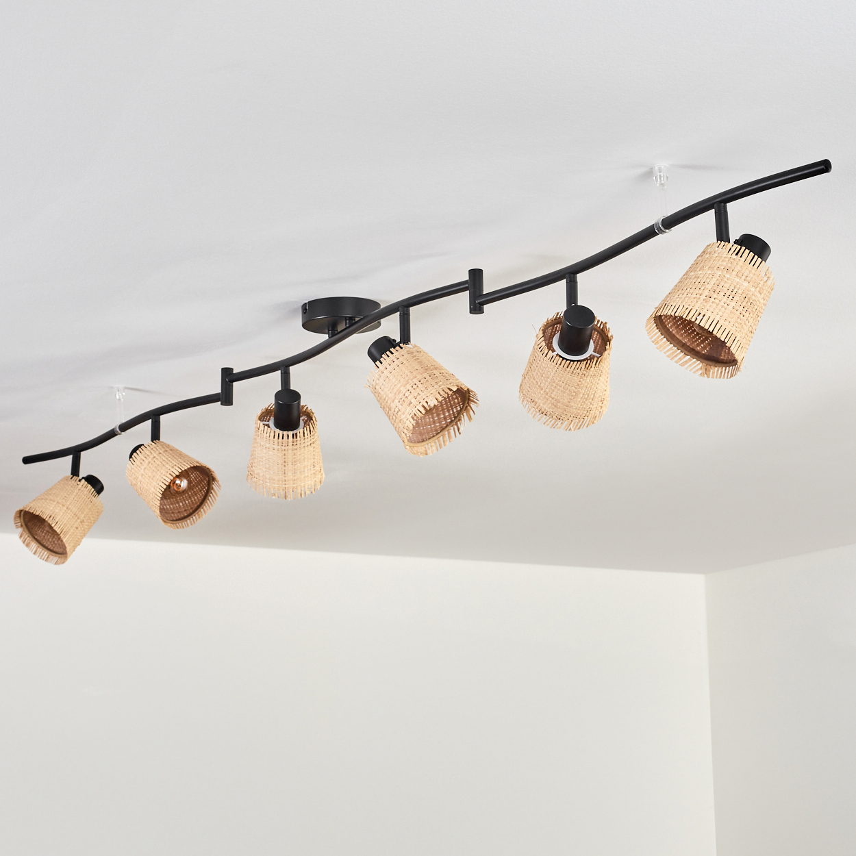 Eine schwarze Schienenbeleuchtung mit sieben gewebten Rattan-Lampenschirmen, die an einer weißen Decke montiert sind. Die Schirme zeigen in verschiedene Richtungen.