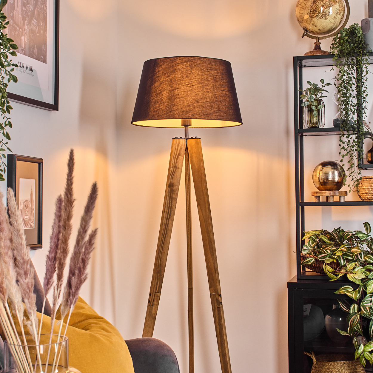 Eine hölzerne Stativ-Stehlampe mit schwarzem Schirm. Rechts ein Regal mit Pflanzen und einem Globus. Links ein Gemälde und Pampasgras.