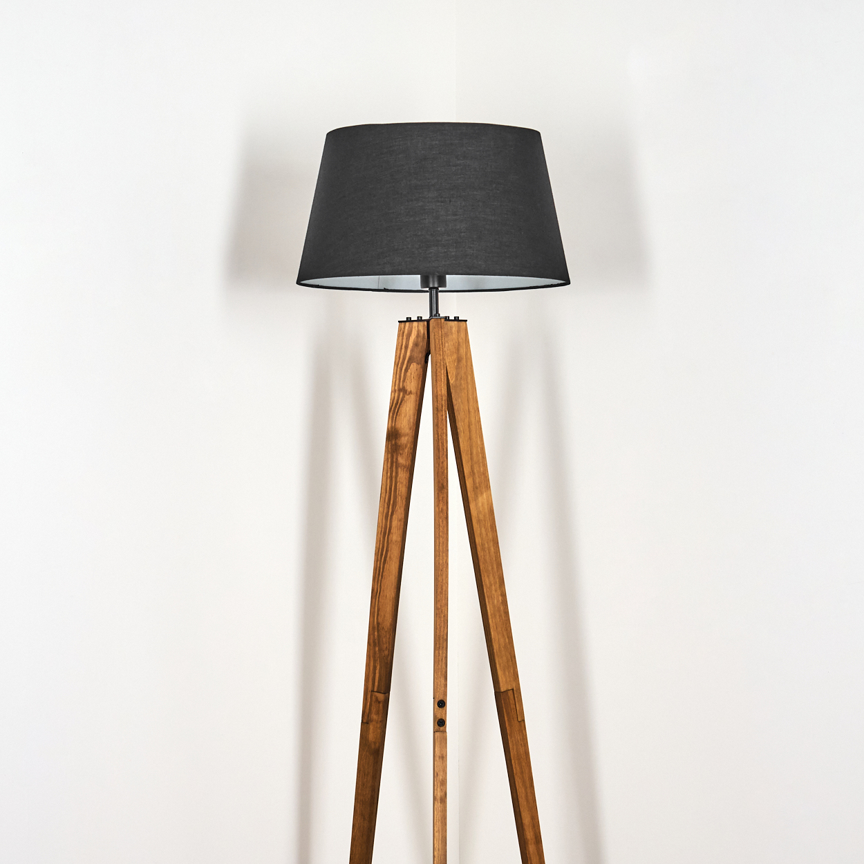 Eine hölzerne Stativ-Stehlampe mit schwarzem Lampenschirm steht vor einer weißen Wand. Die Lampe ist zentriert.