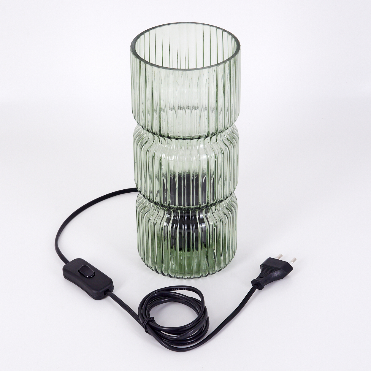 Eine grüne Glaslampe mit schwarzem Kabel und Stecker steht auf einer weißen Oberfläche. Die Lampe ist zylindrisch und gerippt.