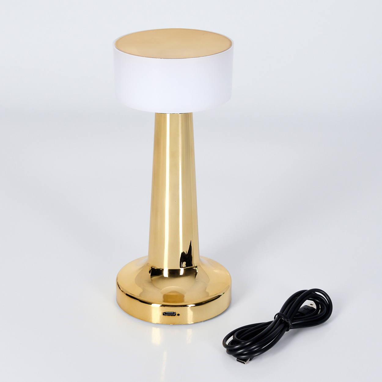 Eine goldene Lampe mit weißem Schirm steht auf einer weißen Oberfläche. Daneben liegt ein schwarzes Kabel. Die Lampe hat eine zylindrische Form.
