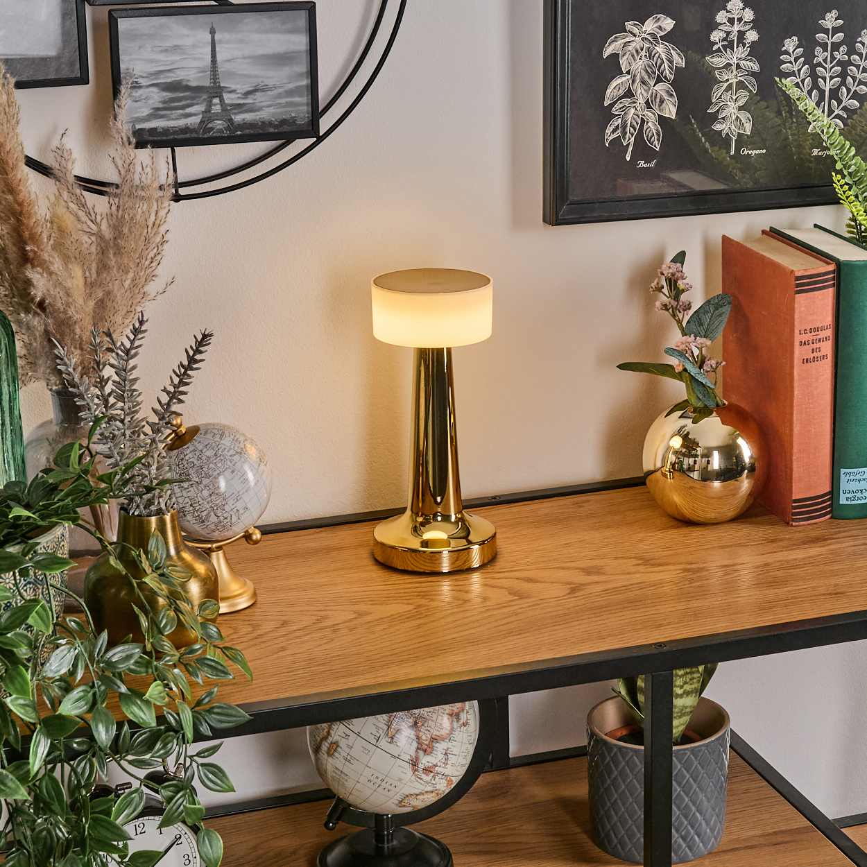 Eine goldene Lampe steht auf einem Holztisch mit Globen, Pflanzen und Büchern. Ein Schwarz-Weiß-Eiffelturm-Bild hängt an der Wand.