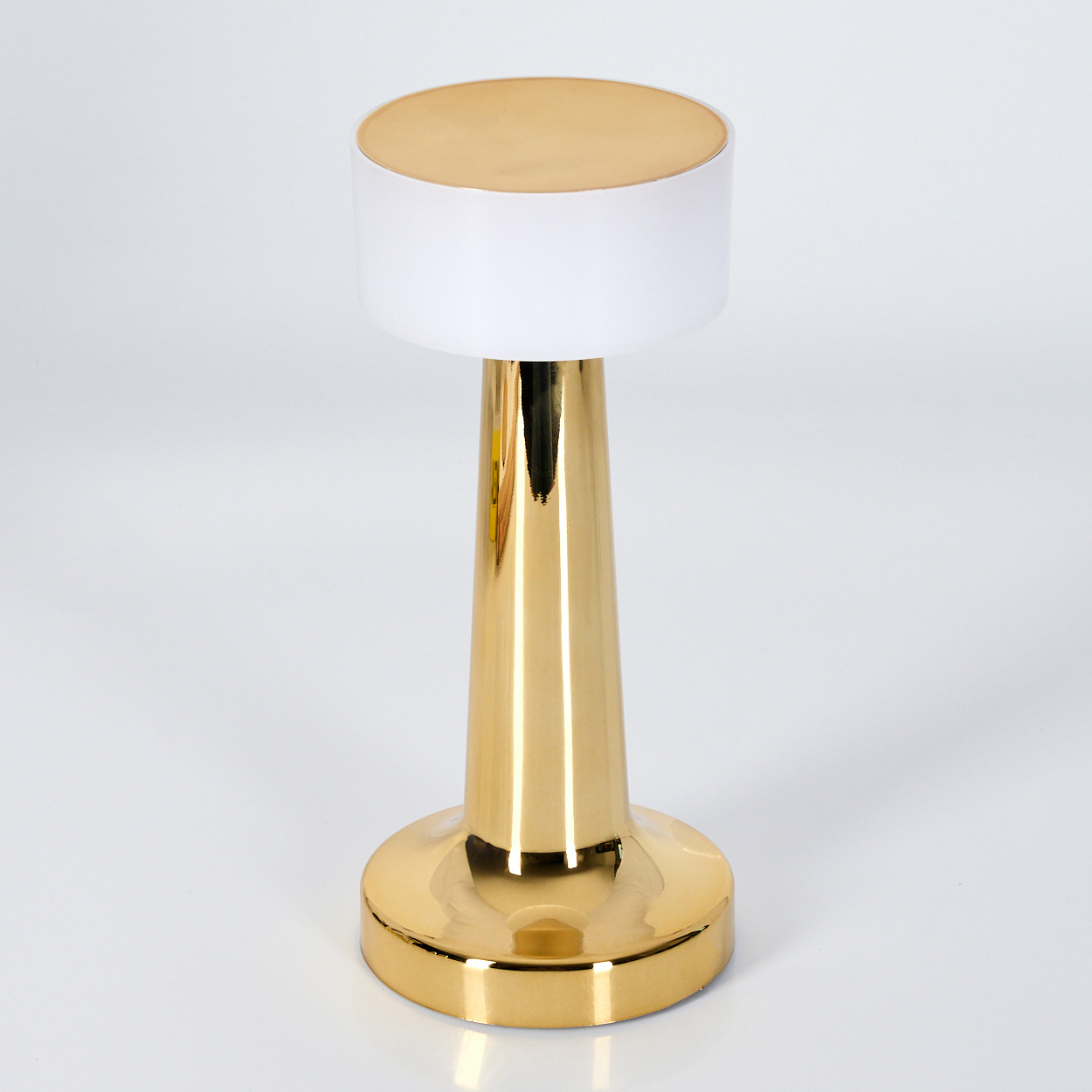 Eine goldene Lampe mit weißem Schirm, auf weißer Oberfläche. Die Lampe hat einen goldenen Sockel, Stiel und Deckel.