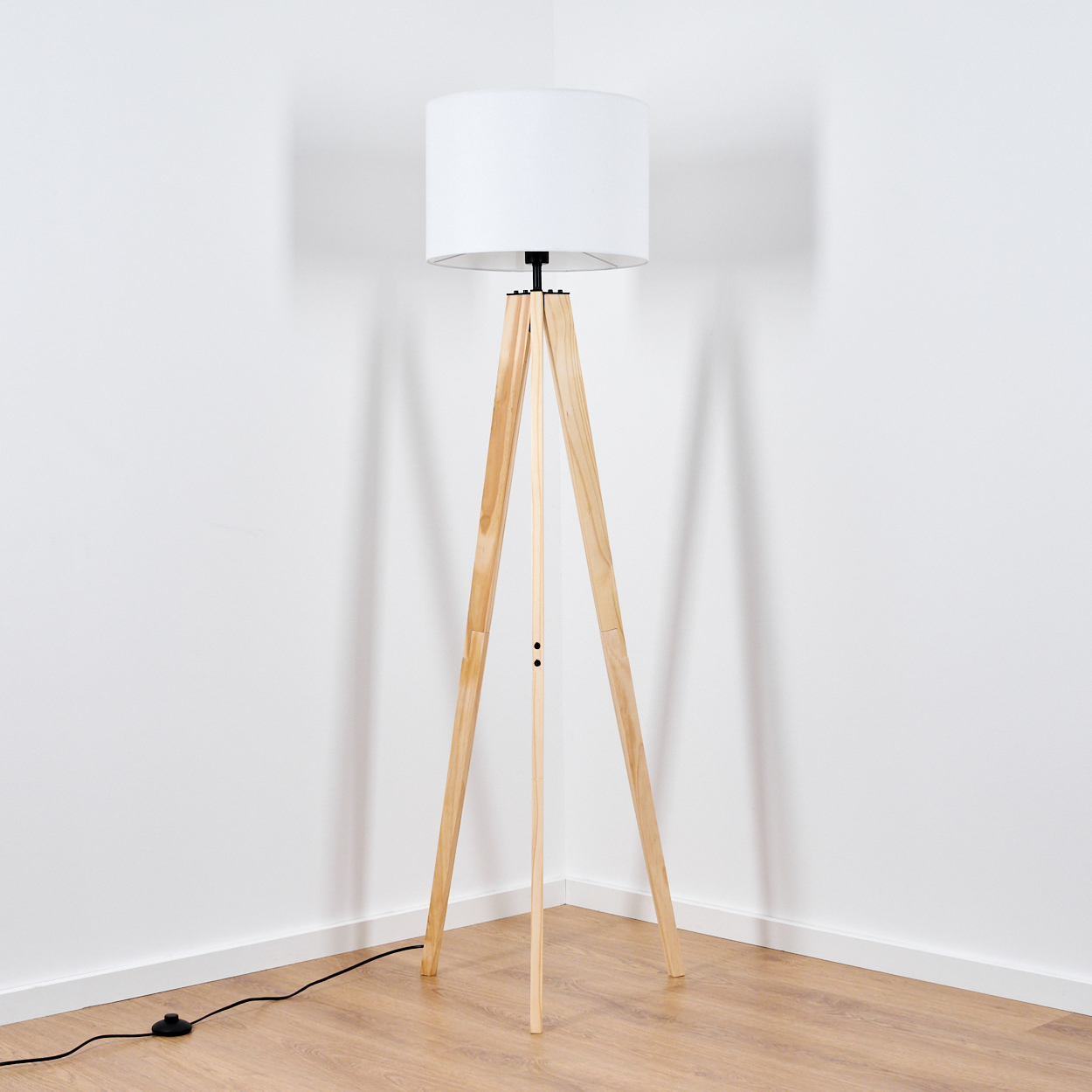Eine hölzerne Stehlampe mit weißem Lampenschirm steht in einer Zimmerecke. Der Boden ist aus Holz und die Wände sind weiß.