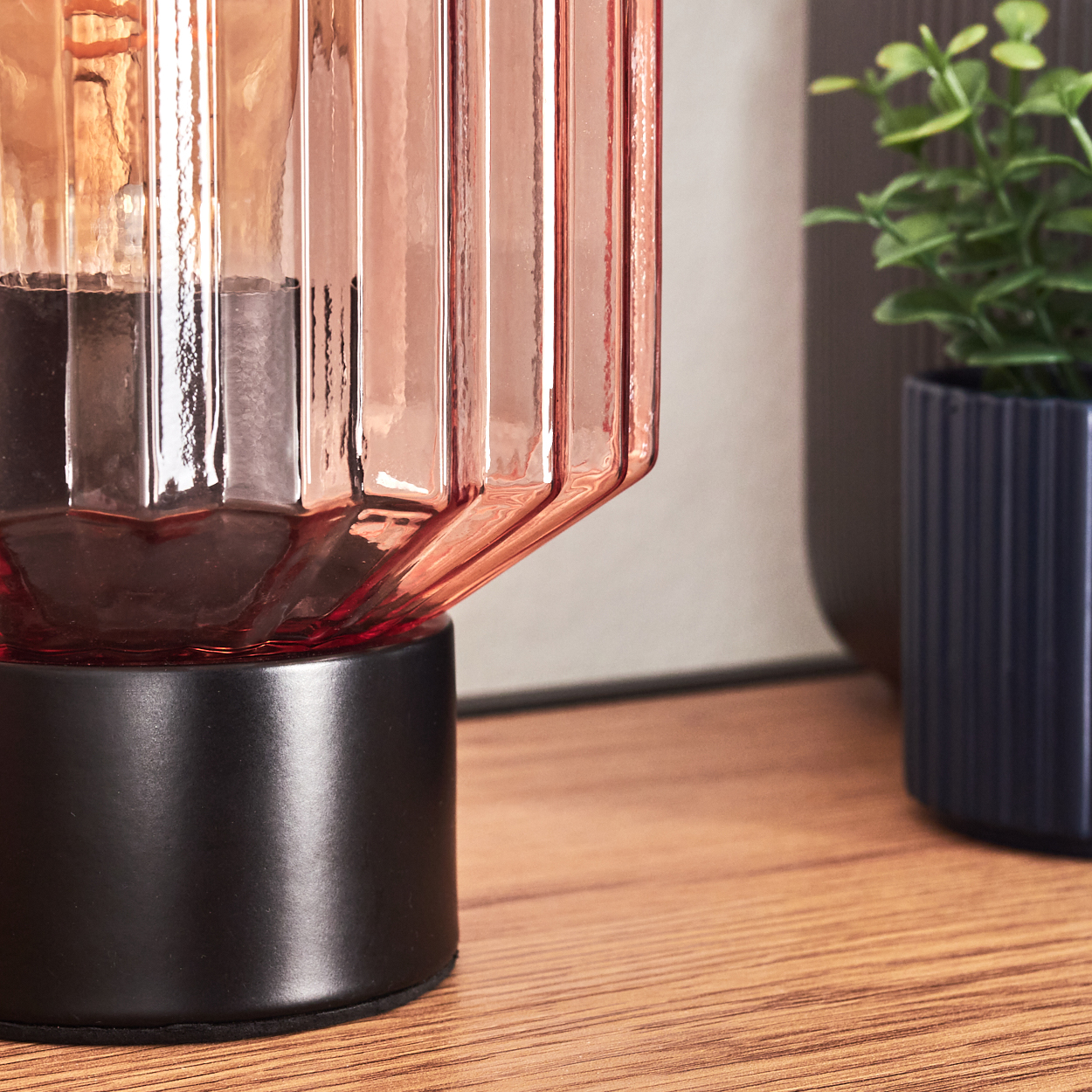 Nahaufnahme einer dekorativen Lampe mit rosafarbenem Glasschirm, schwarzem Sockel und einer kleinen Topfpflanze auf Holzoberfläche.