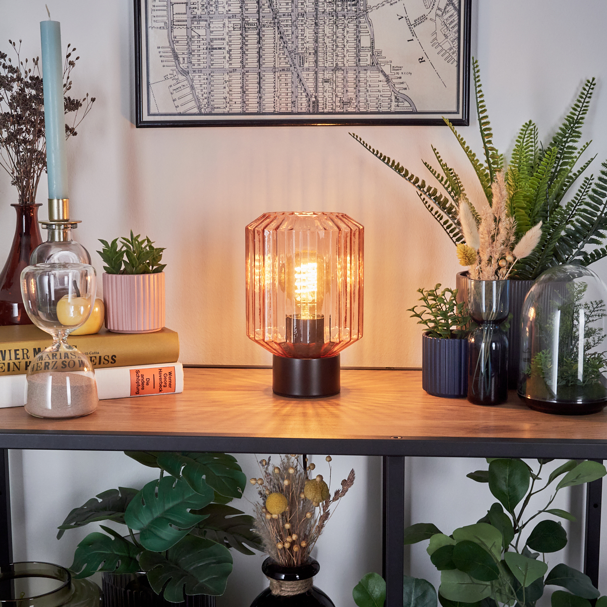 Eine Tischlampe mit rosafarbenem Glasschirm. Verschiedene Pflanzen, Kerzen und Bücher stehen auf einem Holzregal an der Wand.