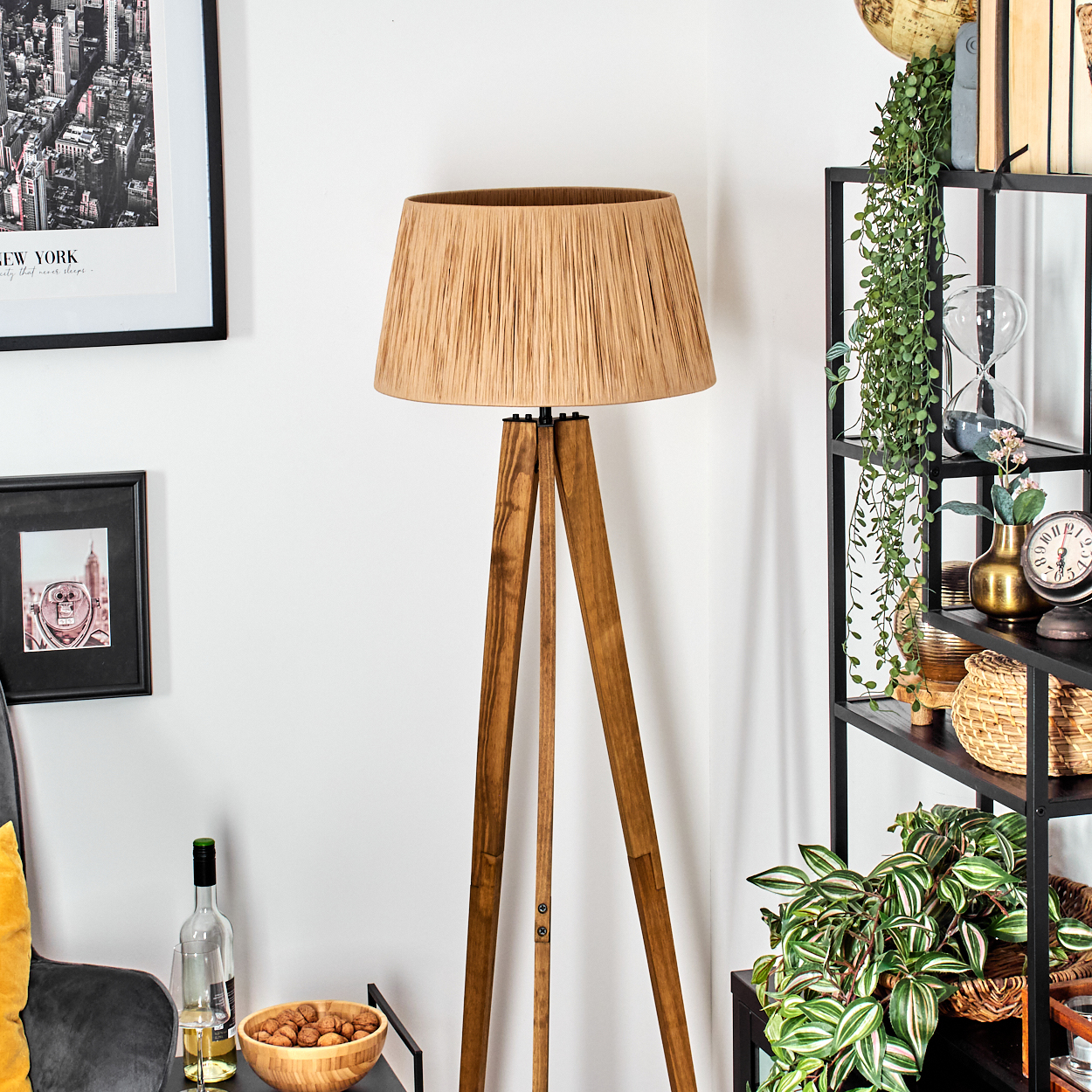 Eine hölzerne Stativ-Stehlampe mit beigem Lampenschirm steht in einem Raum. Regale mit Pflanzen und Dekoration befinden sich rechts.