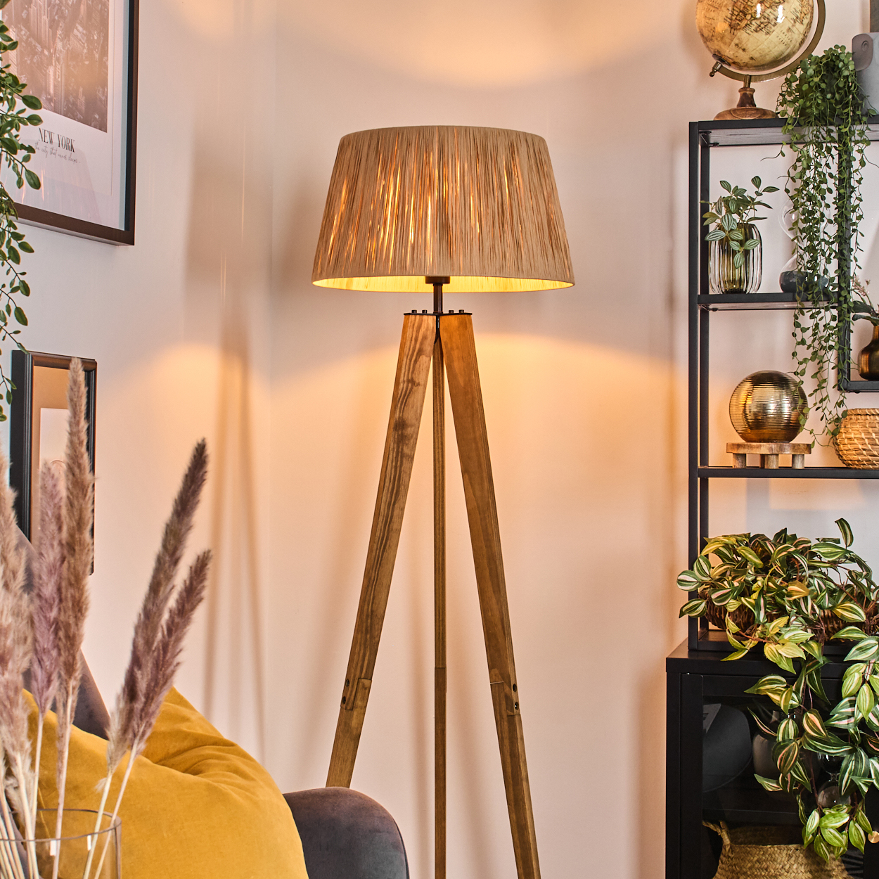Eine Holz-Tripod-Lampe mit beigem Lampenschirm steht in einem Raum. Regale mit Pflanzen sind rechts. Ein gelbes Kissen ist sichtbar.