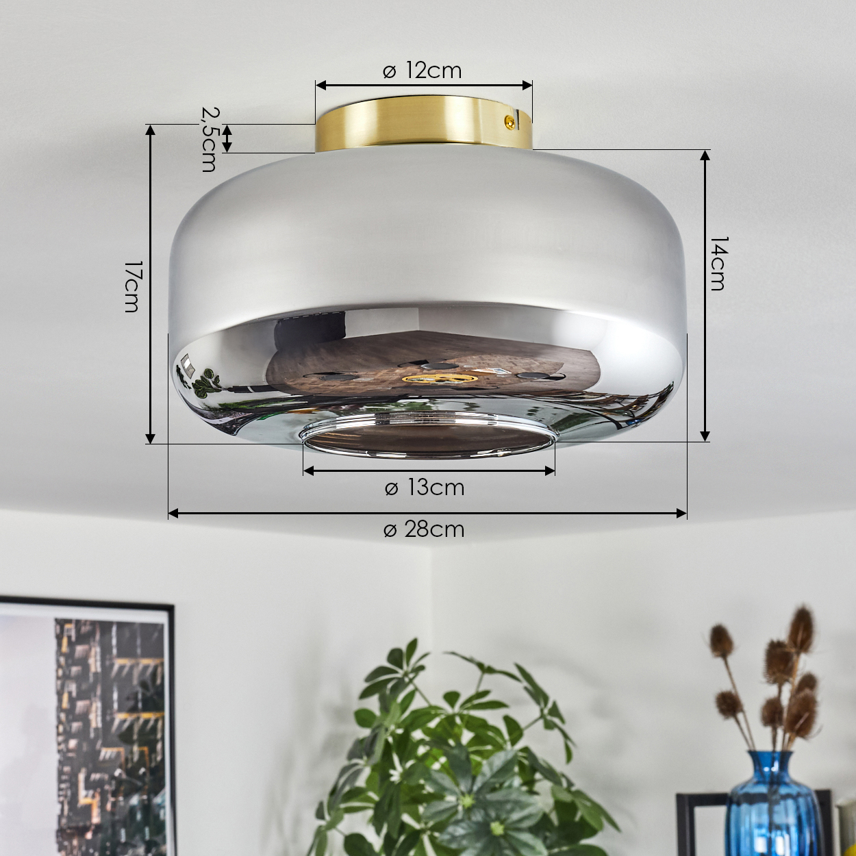 Eine Deckenlampe wird gezeigt. Sie hat einen goldenen Sockel und einen silbernen Schirm, mit angegebenen Abmessungen. Im Hintergrund sind Pflanzen und ein Bild zu sehen.
