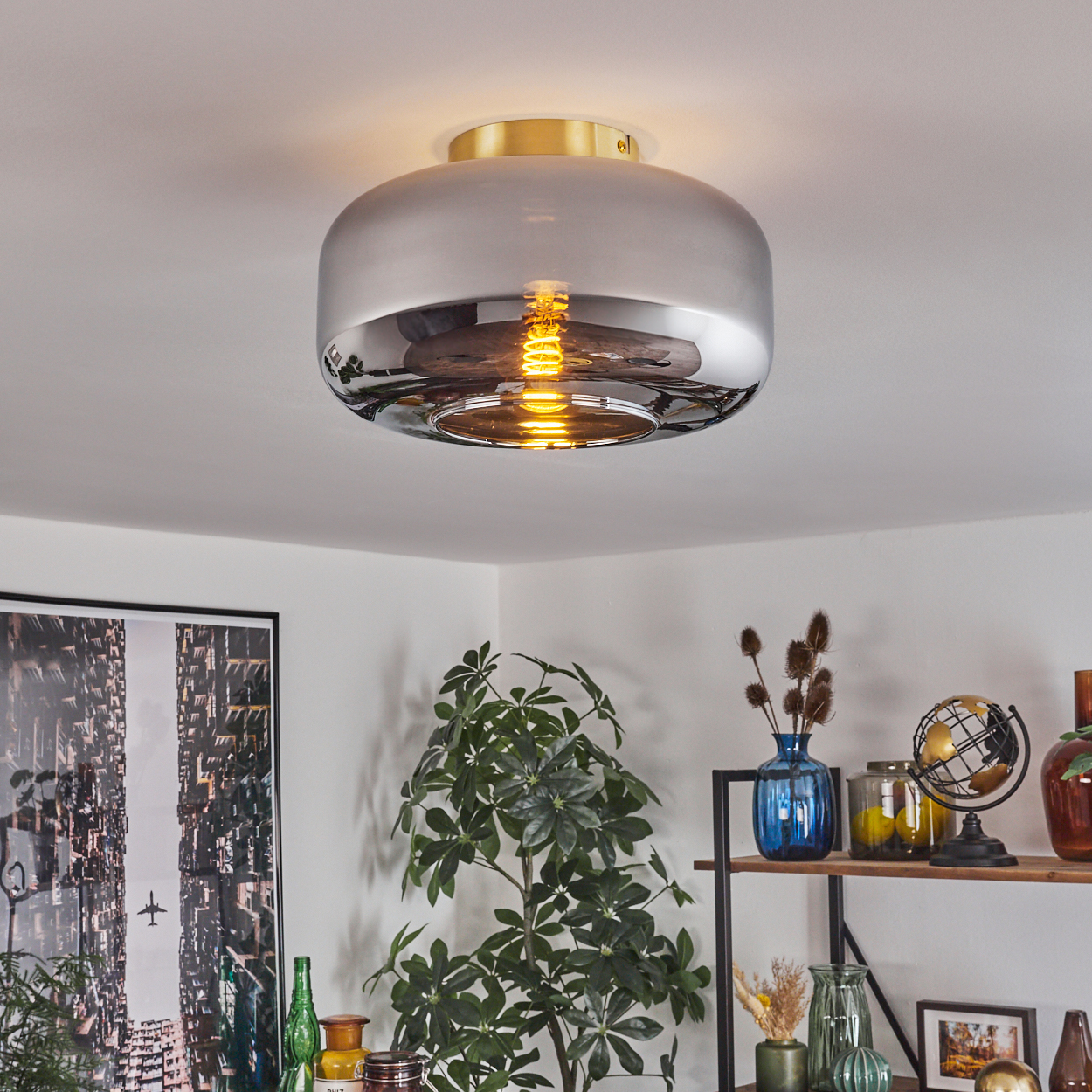 Eine moderne Deckenlampe mit goldfarbenem Sockel und silberfarbenem Schirm. Pflanzen und Regale mit Dekorationen darunter.
