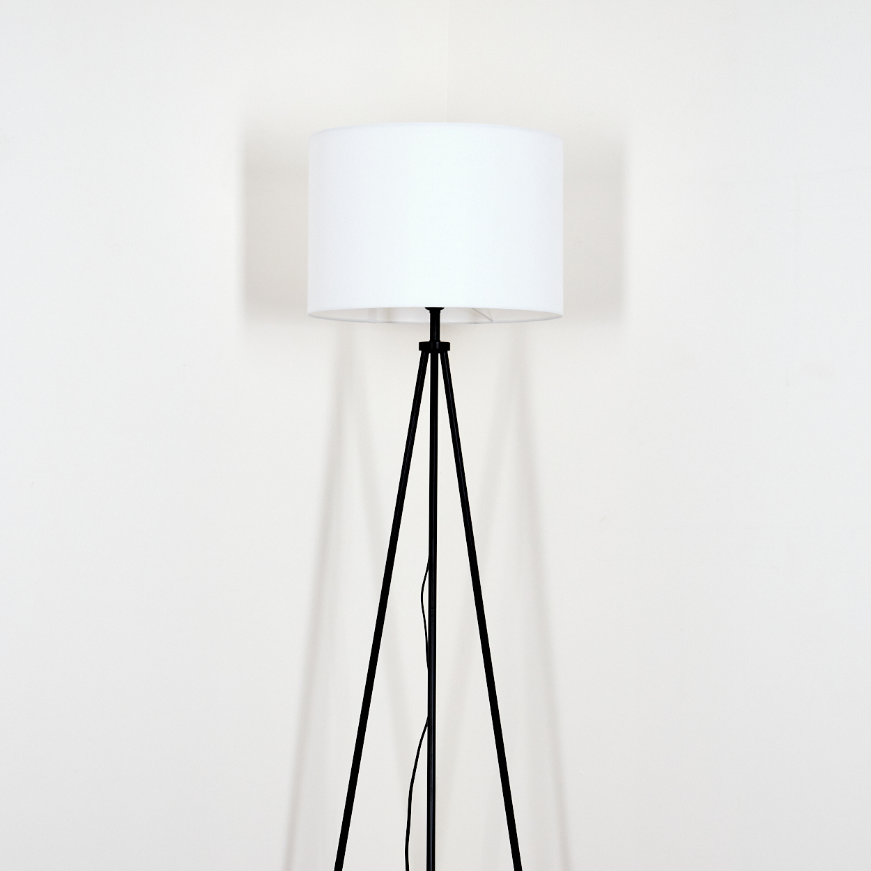 Eine schwarze Stehlampe mit Stativ und einem weißen zylindrischen Lampenschirm steht vor einer weißen Wand.