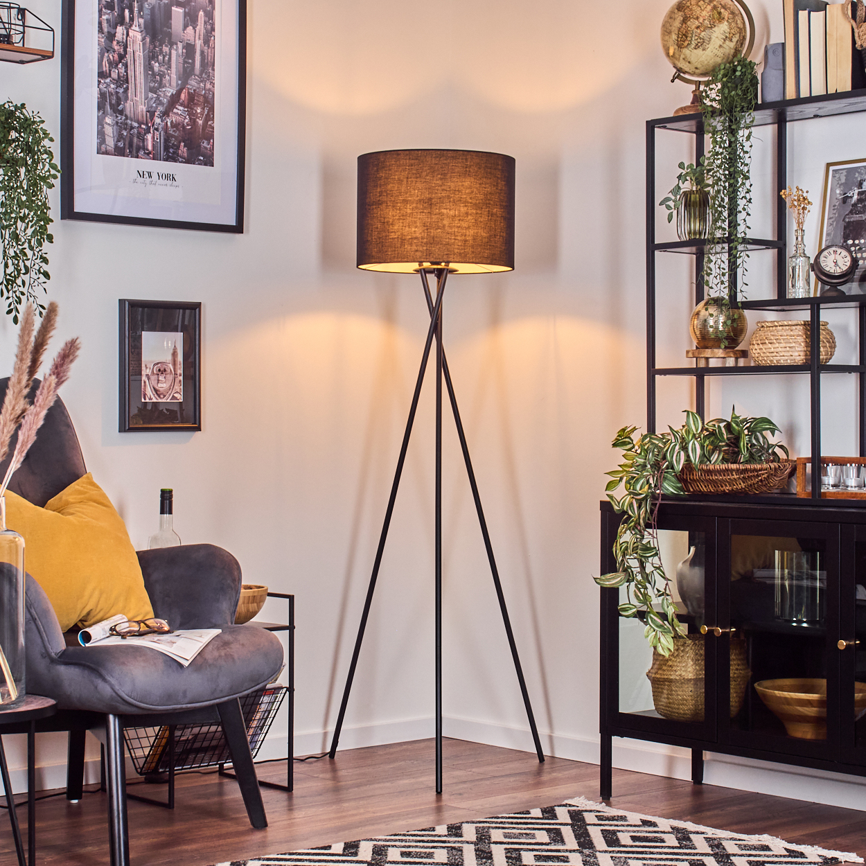 Ein Raum mit einer schwarzen Stehlampe, einem grauen Sessel, einem schwarzen Regal mit Pflanzen und einem gerahmten New York-Stadtbild.