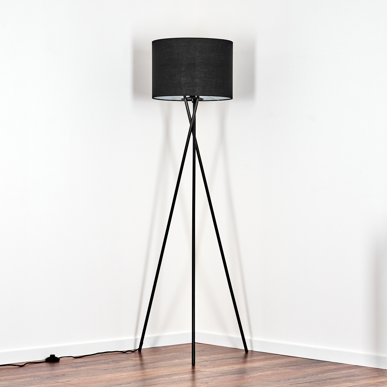 Eine schwarze Stehlampe steht in einer Ecke vor einer weißen Wand mit Holzboden. Die Lampe hat einen zylindrischen Schirm.