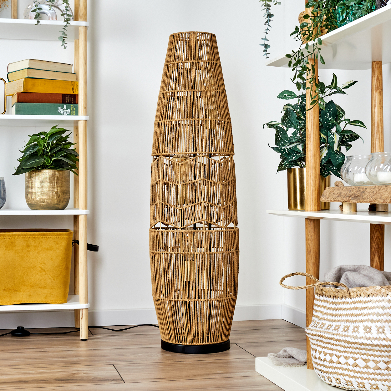 Eine hohe, gewebte Rattanlampe steht im Zentrum. Regale mit Pflanzen, Büchern und Dekor flankieren sie. Ein geflochtener Korb steht auf dem Boden.