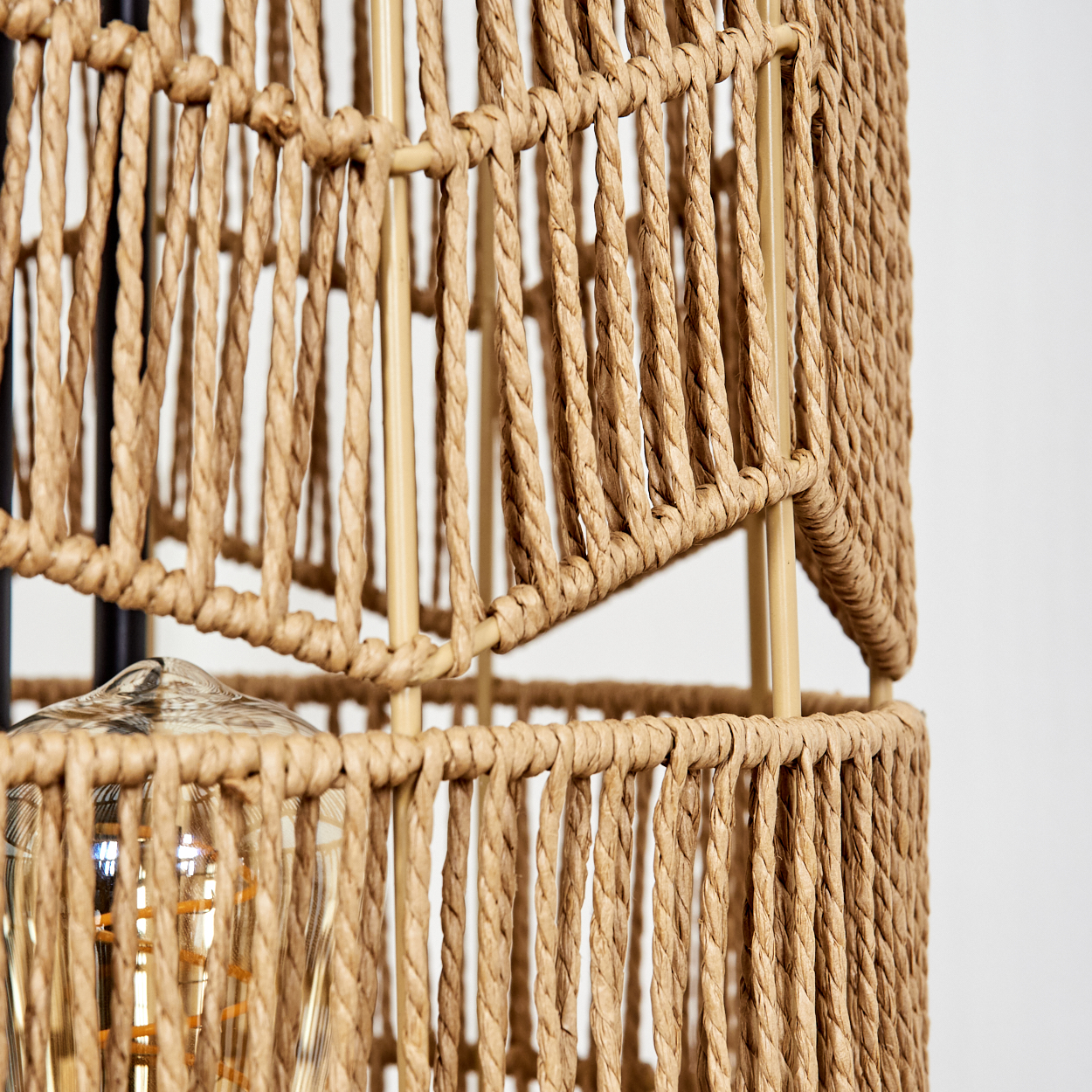 Nahaufnahme eines gewebten Lampenschirms mit Glühbirne. Der Schirm besteht aus braunem, seilähnlichem Material mit goldenem Metallrahmen vor weißem Hintergrund.