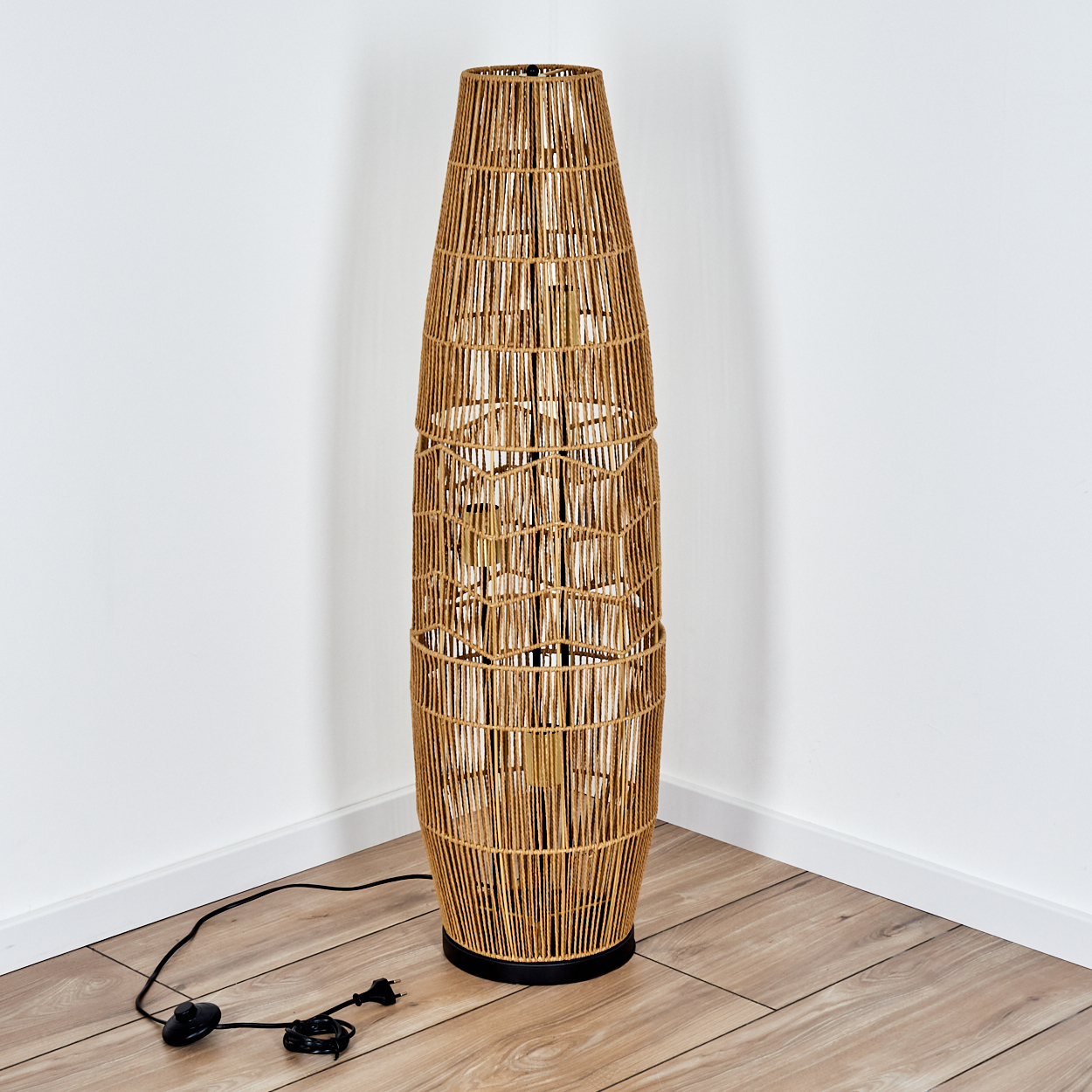 Eine hohe Stehlampe aus geflochtenem Rattan steht in einer Ecke. Sie hat einen schwarzen Sockel und ein schwarzes Kabel. Der Boden ist Holz, die Wand weiß.