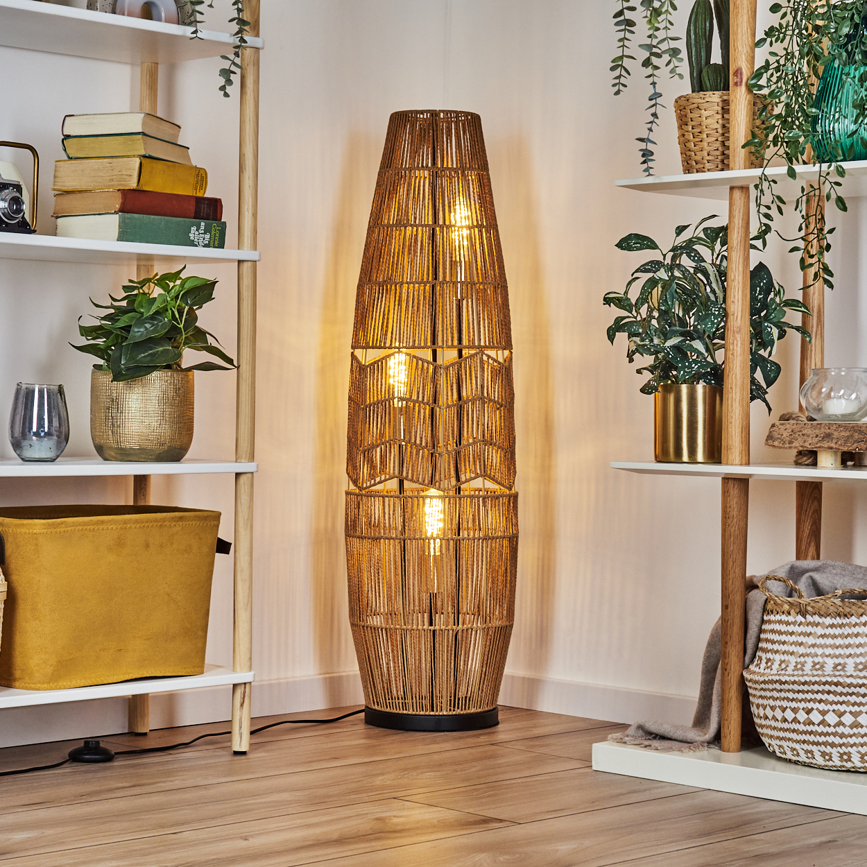Eine gewebte Stehlampe steht zwischen Bücherregalen mit Pflanzen und Dekoration. Die Lampe strahlt warmes Licht aus, das die Ecke des Raumes erhellt.