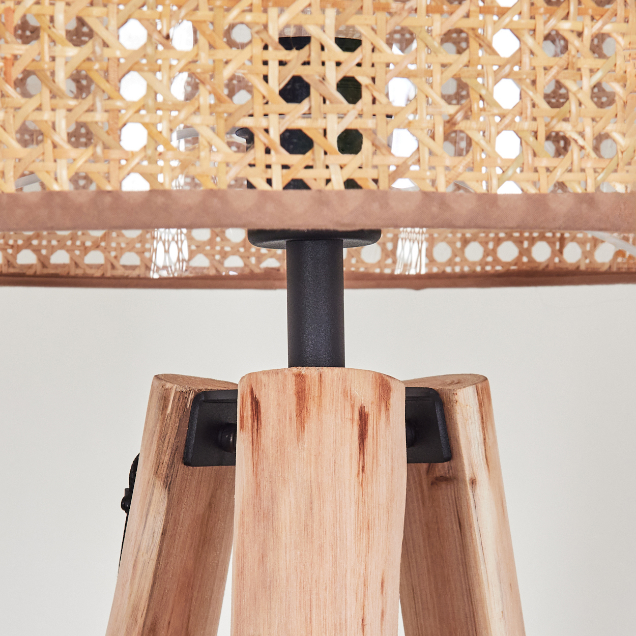 Nahaufnahme einer hölzernen Stativlampe. Der Lampenschirm ist aus gewebtem Rattan. Die Beine sind aus hellem Holz. Eine schwarze Metallsäule hält den Schirm.