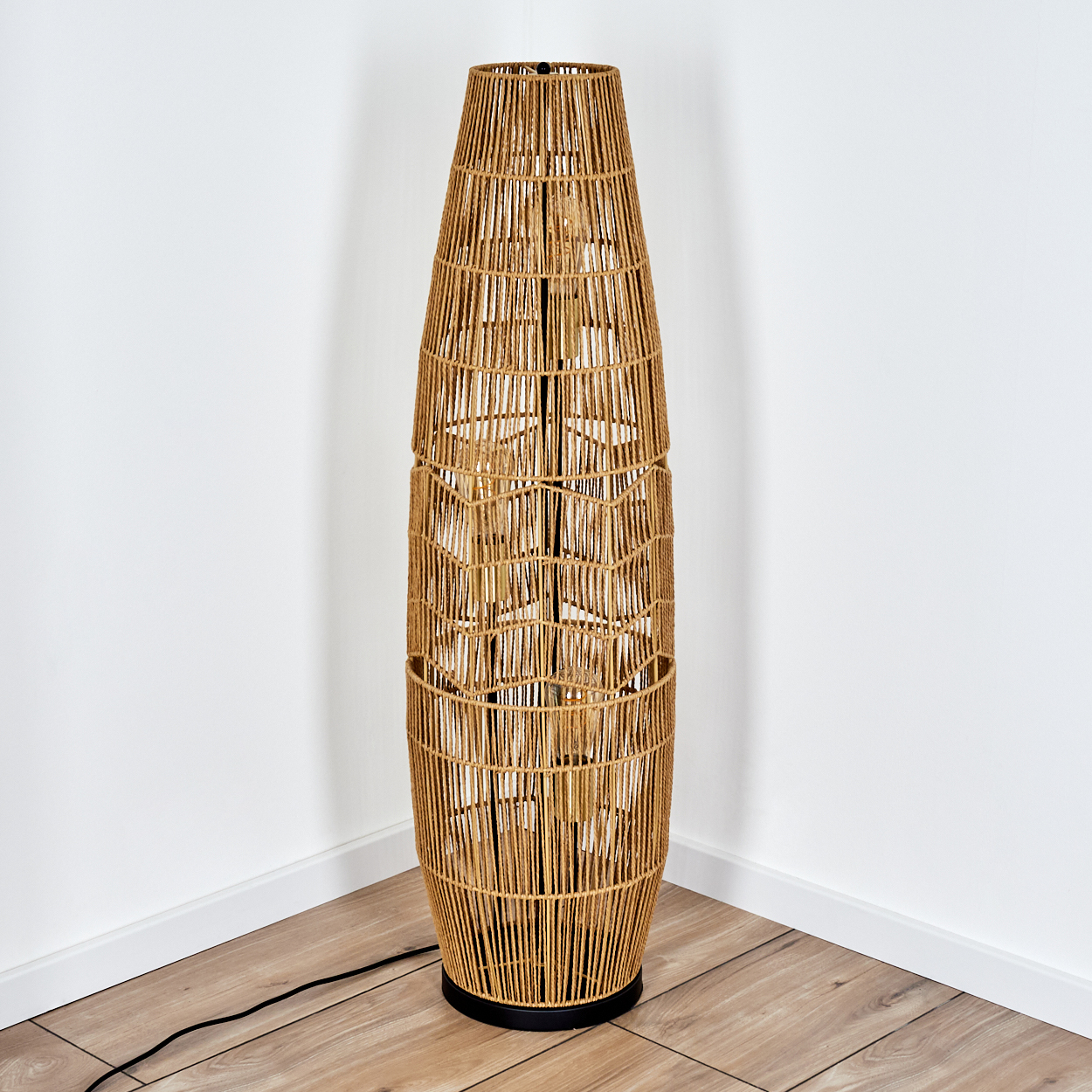 Eine hohe Stehlampe aus geflochtenem Weidengeflecht steht in einer Ecke vor weißen Wänden und einem Holzboden. Sie hat einen schwarzen Sockel.