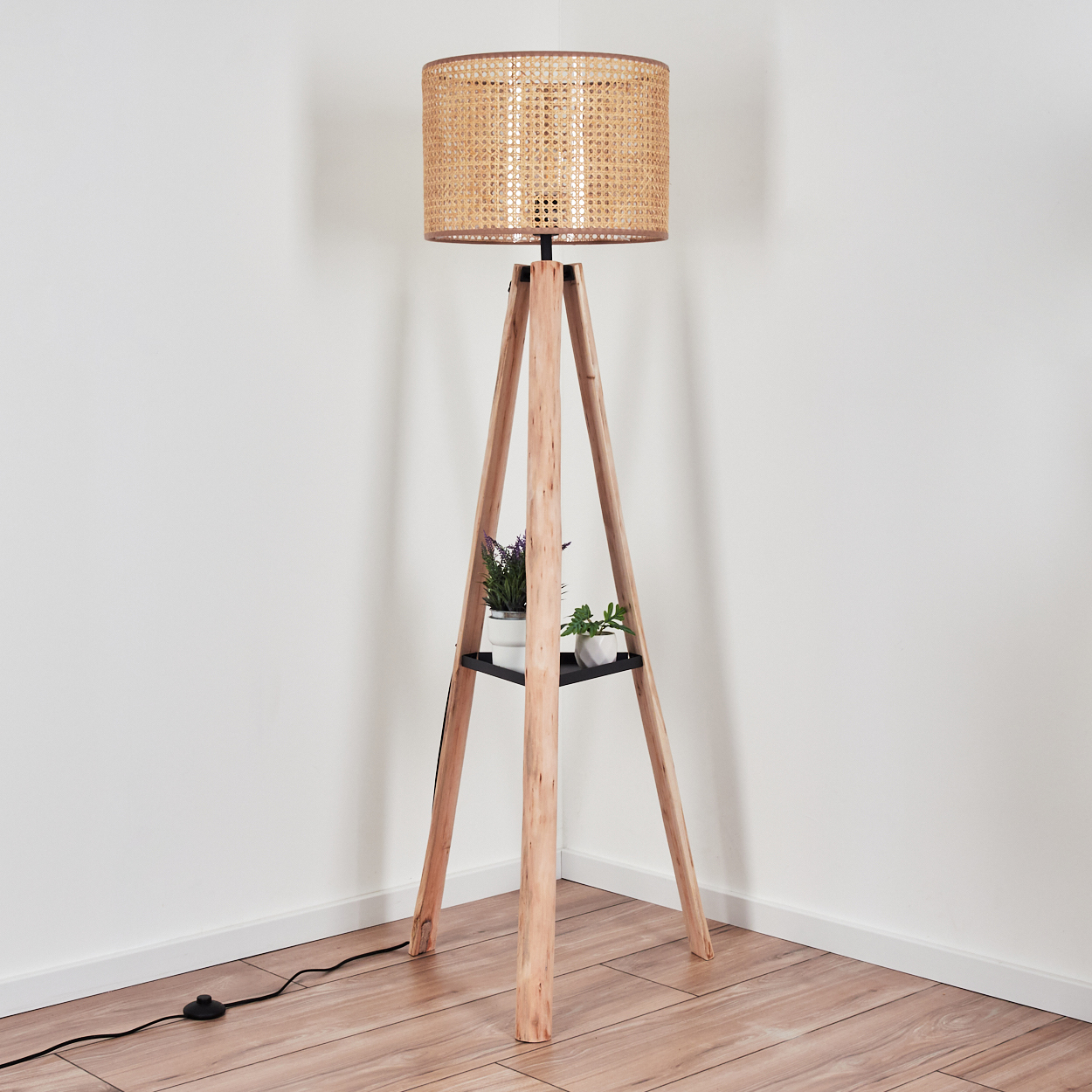 Eine hölzerne Stehlampe mit gewebtem Schirm. Sie hat ein Regal mit Pflanzen. Die Lampe steht auf einem Holzboden in einem weißen Raum.