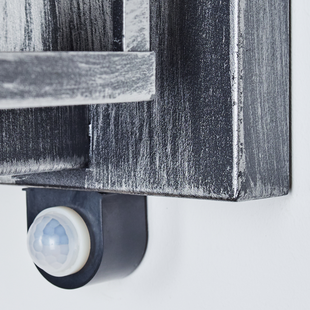 Nahaufnahme einer metallischen, strukturierten Box mit einem Bewegungssensor. Die Box ist silber und schwarz, an einer weißen Wand befestigt.
