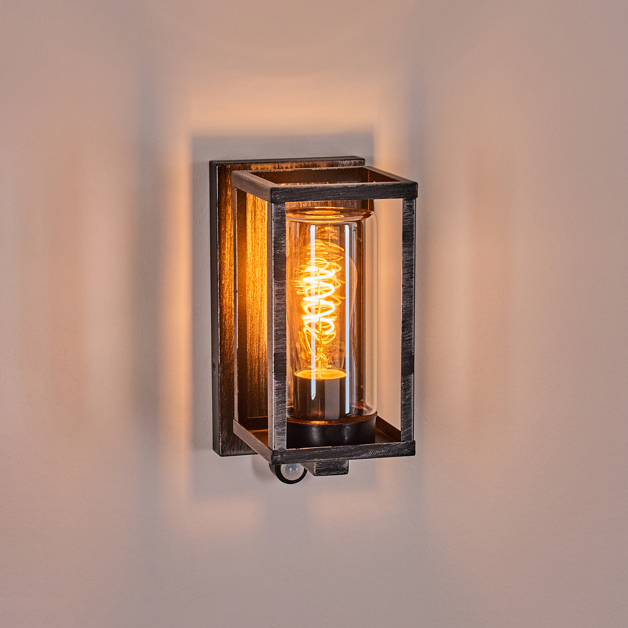 Wandleuchte im Vintage-Design. Sie hat einen rechteckigen Metallrahmen, ein Glasrohr mit einer Wendelglühbirne und ist an einer Wand befestigt.
