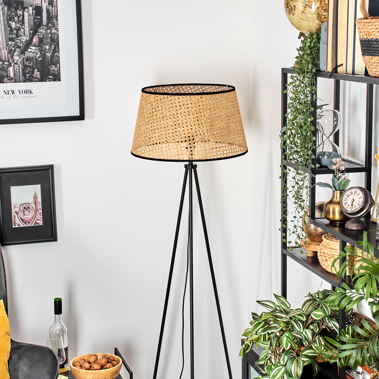 Eine Stehlampe mit gewebtem Schirm steht neben einem Regal mit Pflanzen und Dekoration. Ein New York-Druck hängt an der Wand.