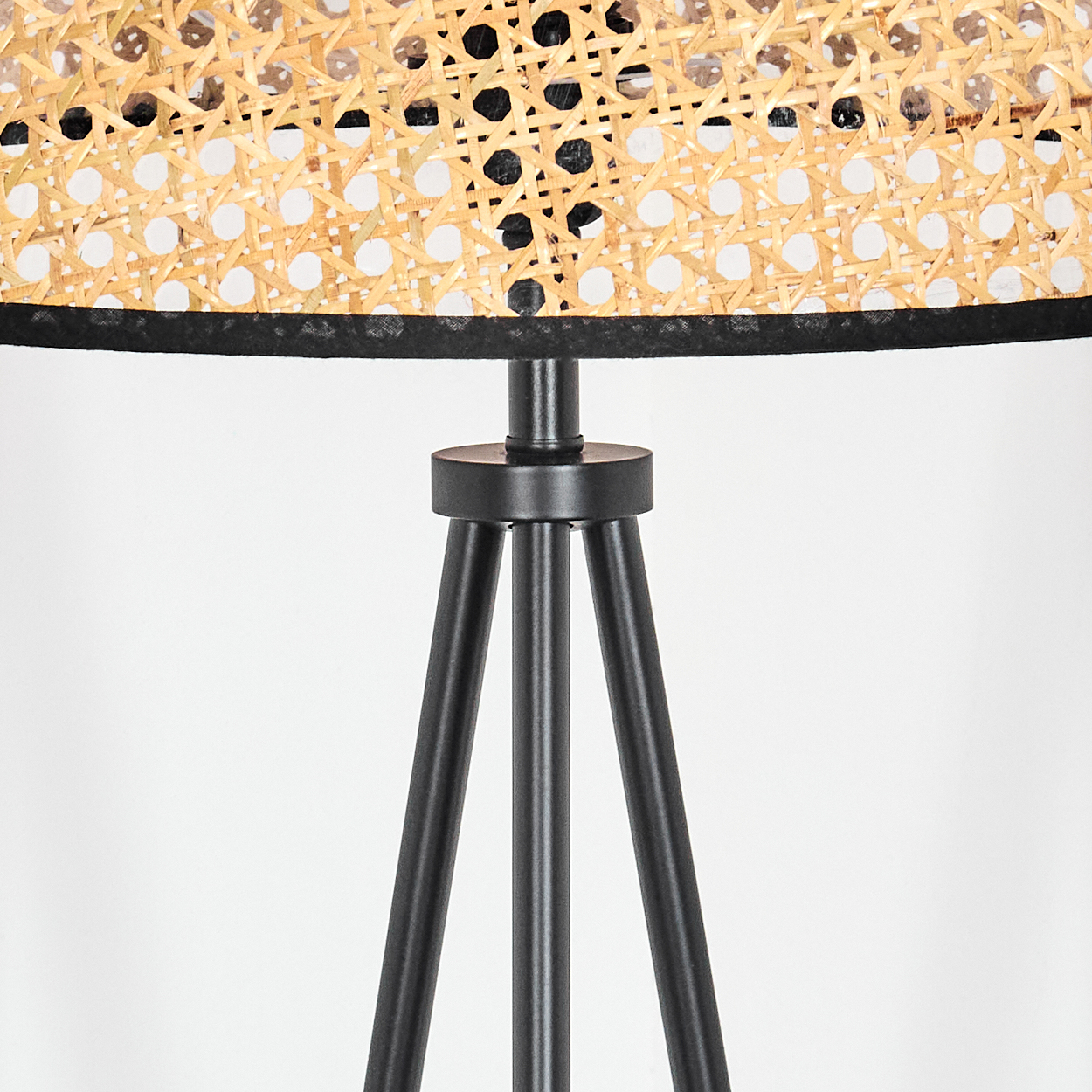 Nahaufnahme einer Lampe. Sie hat einen Lampenschirm aus geflochtenem Rattan mit schwarzem Rand, der von schwarzen Metall-Dreibein-Beinen getragen wird.