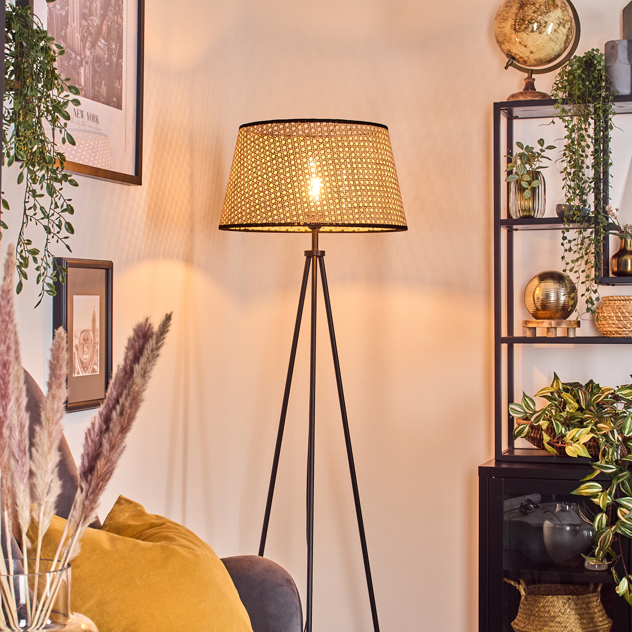 Eine beleuchtete Stativlampe mit gewebtem Schirm steht in einem Raum mit Pflanzen, Kunst und einem Regal.