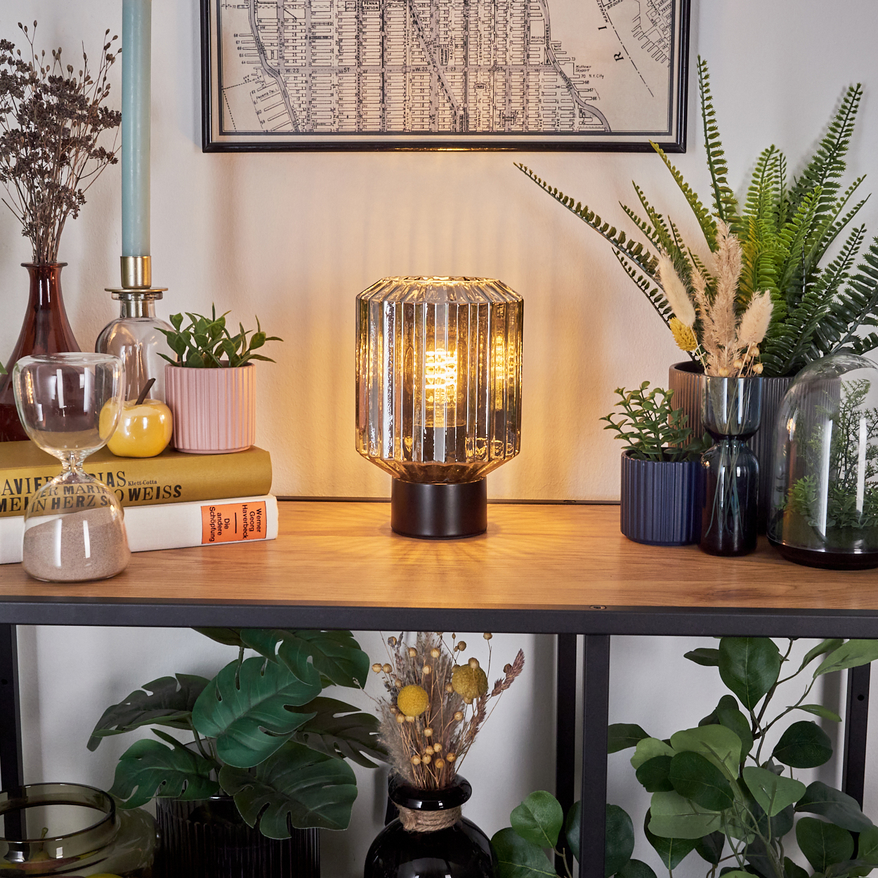 Eine beleuchtete Lampe steht auf einem Regal mit Pflanzen und Büchern. Eine Karte hängt über dem Regal.