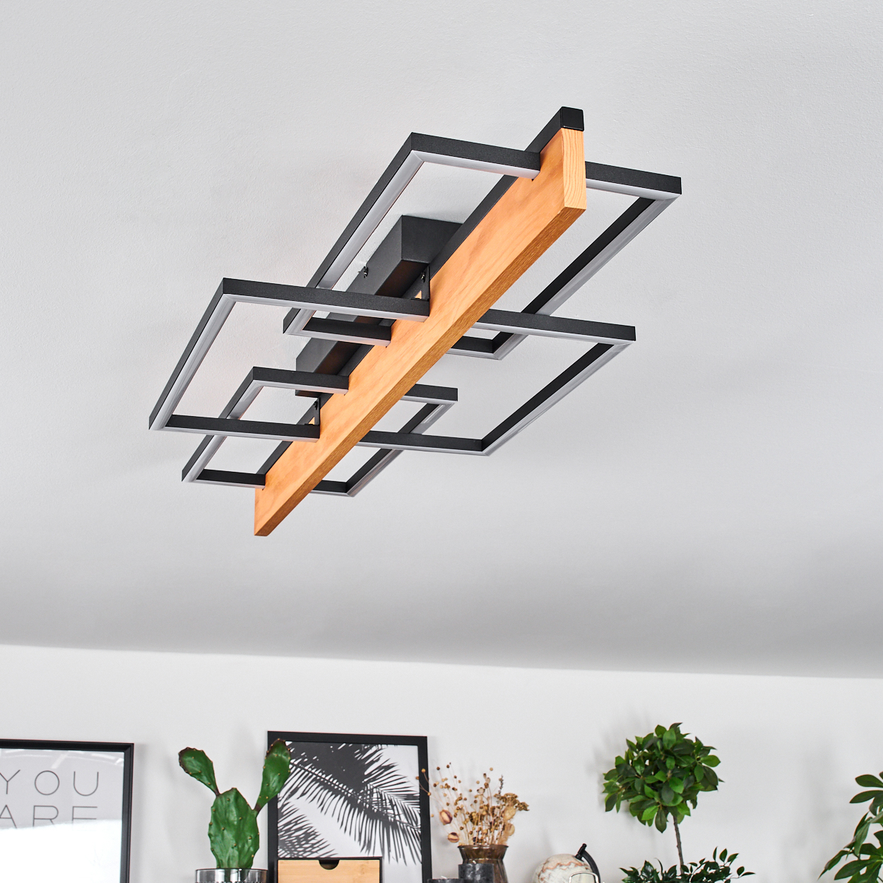 Moderne Deckenleuchte. Sie hat einen Holzbalken mit schwarzen geometrischen Formen. Darunter befinden sich gerahmte Bilder und Pflanzen.