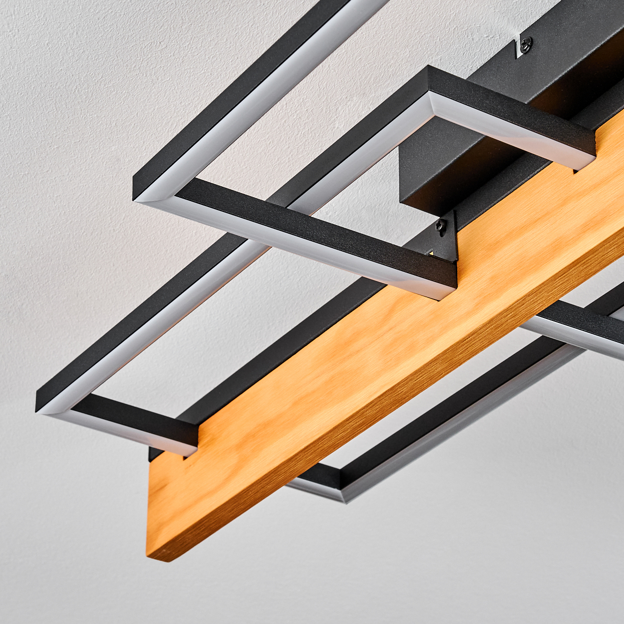 Moderne Deckenleuchte mit schwarzen quadratischen Metallrahmen, weißen Lichtleisten und einem Holzbalken vor weißer Decke.