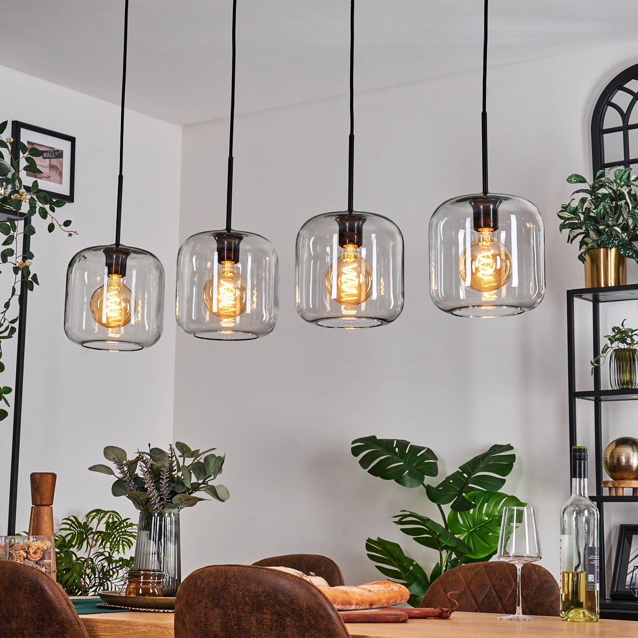 Vier Glas-Pendelleuchten hängen über einem Holztisch. Der Raum hat Pflanzen, Weingläser und ein Regal mit Dekor.