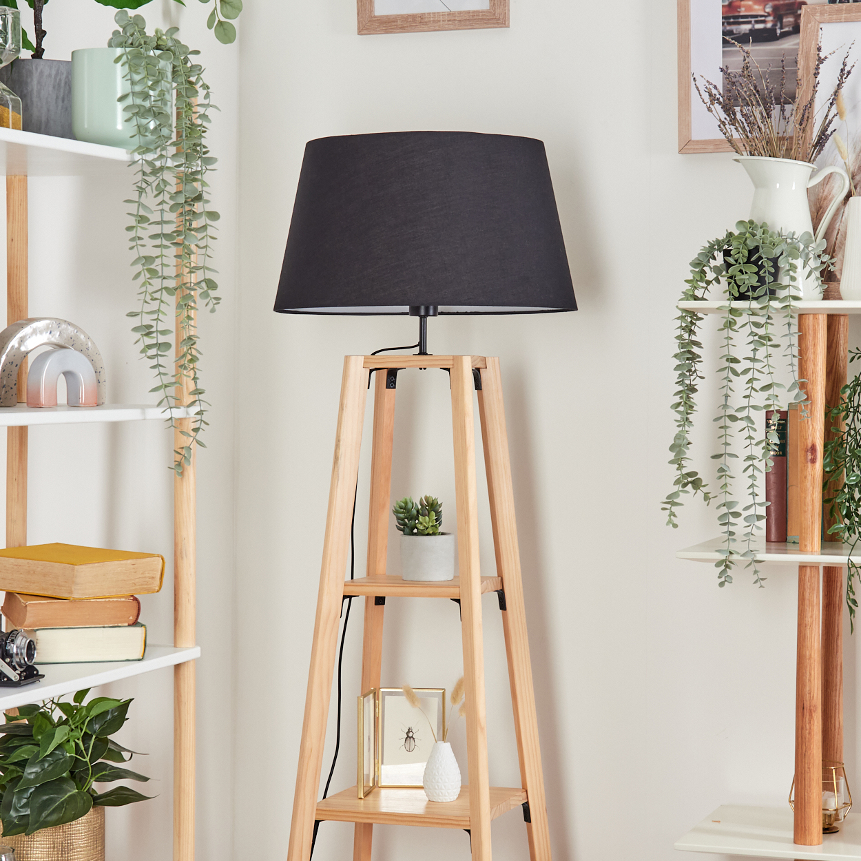 Holz-Stehlampe mit schwarzem Lampenschirm, Regale mit Pflanzen und Dekoration. Hintergrund hat gerahmte Bilder und weiße Wand.