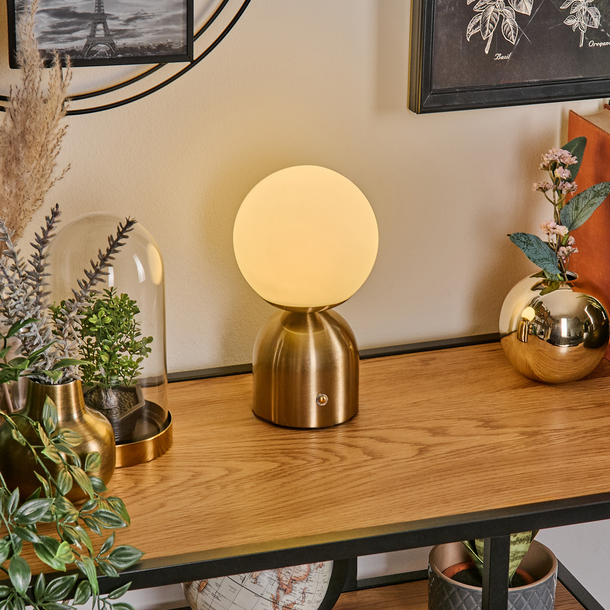 Ein Tisch mit einer Messinglampe, Vasen mit Pflanzen und gerahmten Bildern. Eine Glaskuppel mit einer Pflanze. Holztisch.