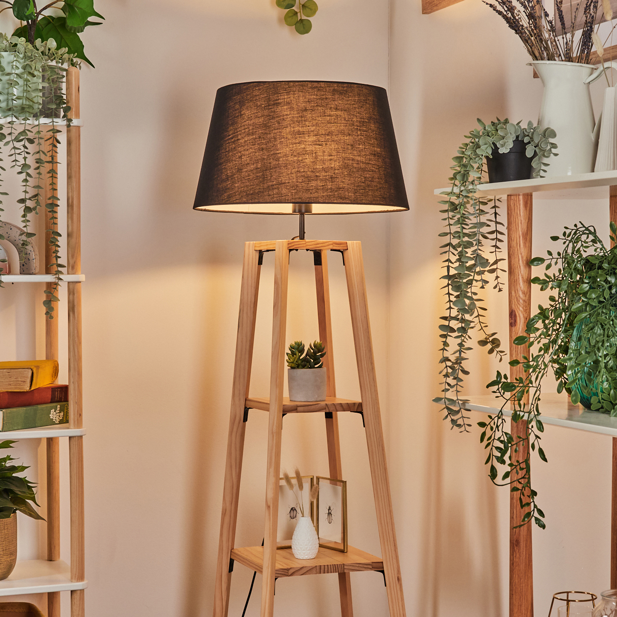 Eine Holzlampe mit schwarzem Lampenschirm steht neben Regalen mit Pflanzen und Büchern. Eine Vase und Rahmen stehen auf den Regalen der Lampe.