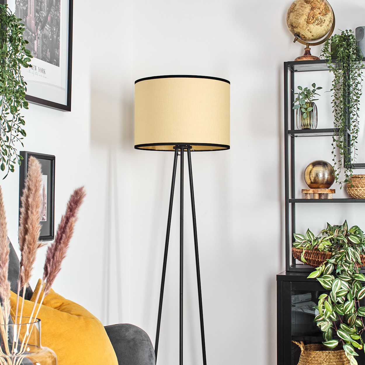 Eine Stehlampe mit beigem Schirm und schwarzen Beinen. Pflanzen, Globus und Regale im Hintergrund.
