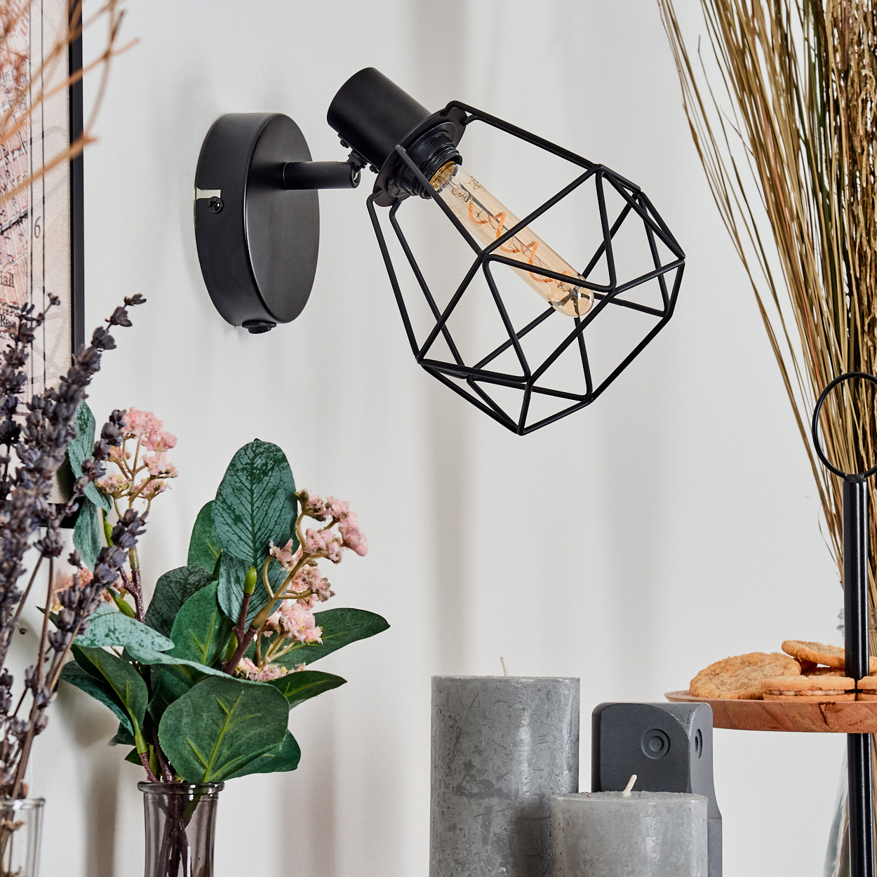 Eine schwarze Wandlampe mit käfigartigem Schirm, neben Blumen, Kerzen und Keksen auf einem Holzständer.