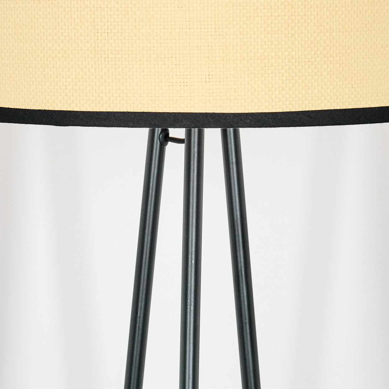 Eine Lampe mit cremefarbenem Schirm und schwarzem Rand. Sie steht auf drei schwarzen Beinen vor weißem Hintergrund.