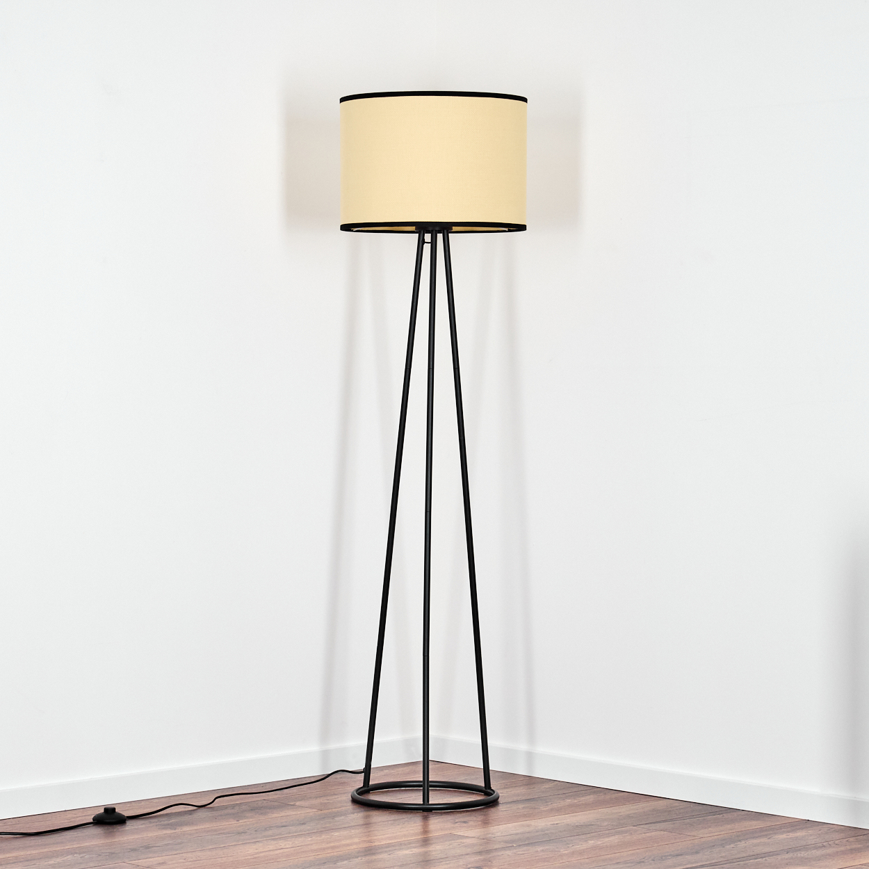 Eine Stehlampe mit cremefarbenem Lampenschirm und schwarzem Rahmen steht in einer Ecke. Der Boden ist aus Holz, die Wände sind weiß.