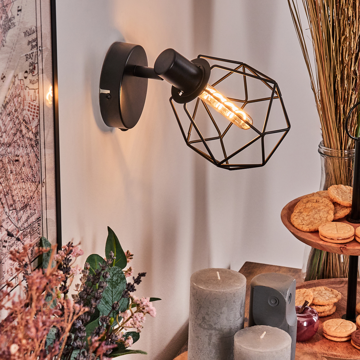 Eine schwarze Wandlampe mit geometrischem Schirm. Kekse, Kerzen und Blumen stehen auf dem Tisch. Im Hintergrund ist eine Karte zu sehen.