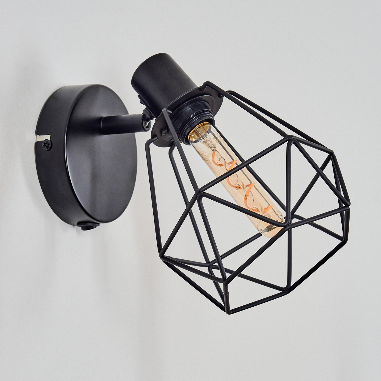 Eine schwarze Wandleuchte mit einem geometrischen Käfig um eine Glühbirne. Sie ist an einer weißen Wand montiert. Die Glühbirne leuchtet.