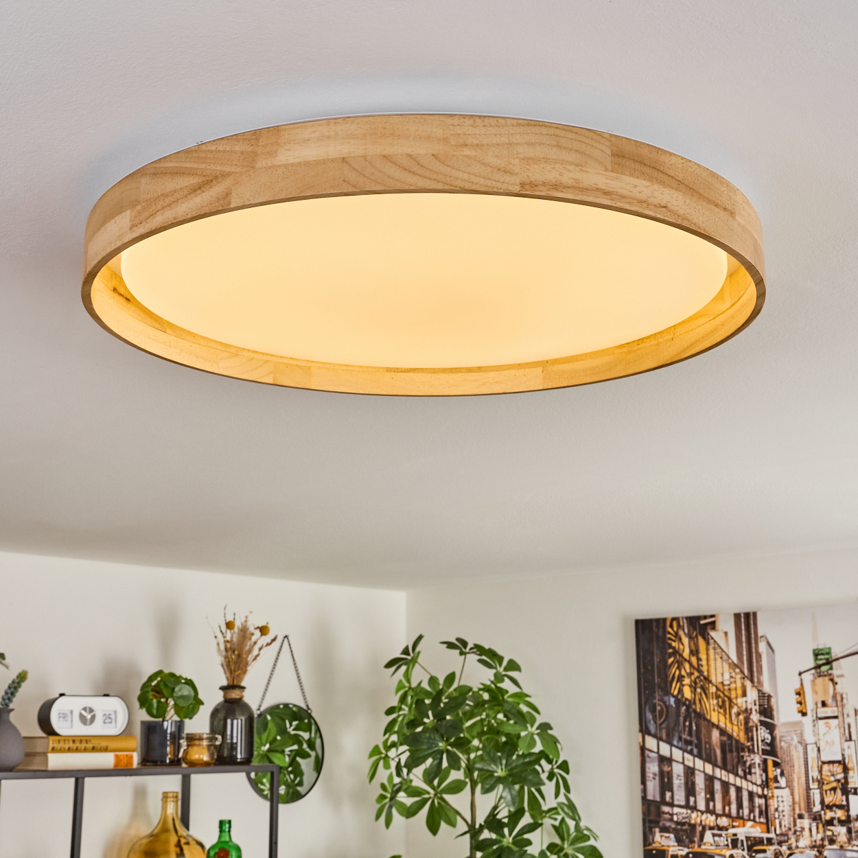 Eine kreisförmige Deckenleuchte aus Holz mit hellem Zentrum. Darunter ein Regal mit Dekorationen und Pflanzen und ein Bild an der Wand.